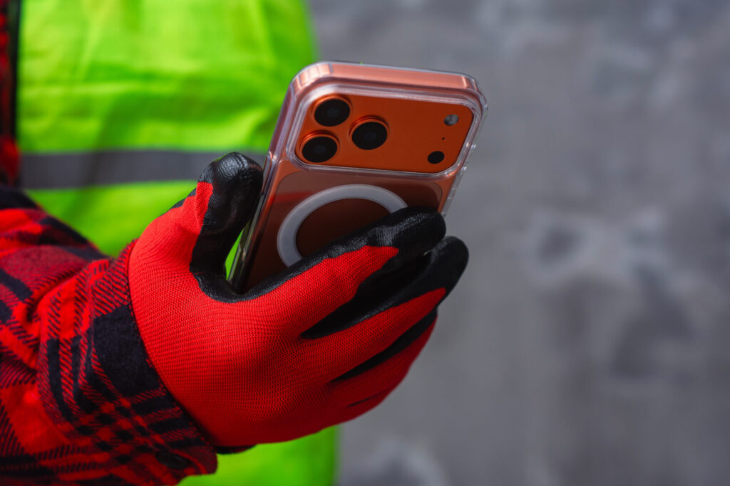Worker hands wearing protective gloves holding smartphone at construction site. Concept of mobile technology in construction industry, digital tools, communication and modern industrial work