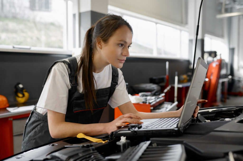 Back view on woman auto engineer doing computer diagnostic using laptop computer for finding car failure causes. Automobile inspection at professional auto service workshop