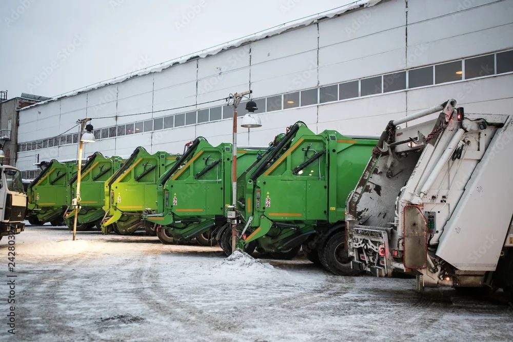 Row of green garbage trucks in a snowy lot.
